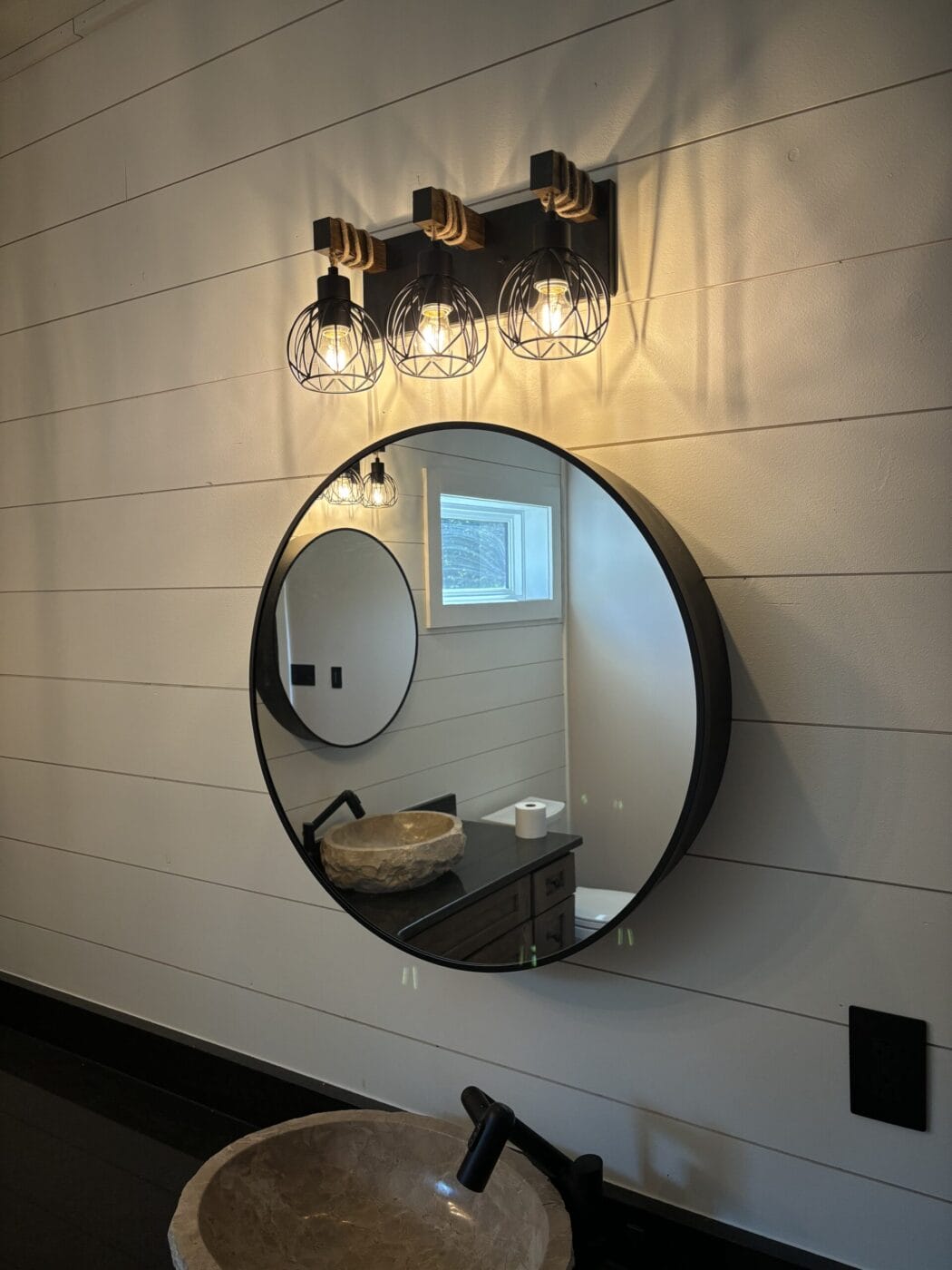 A circular mirror with a stone basin sits below a sconce with three exposed bulb lights on a shiplap wall. A small window is visible in the reflection.
