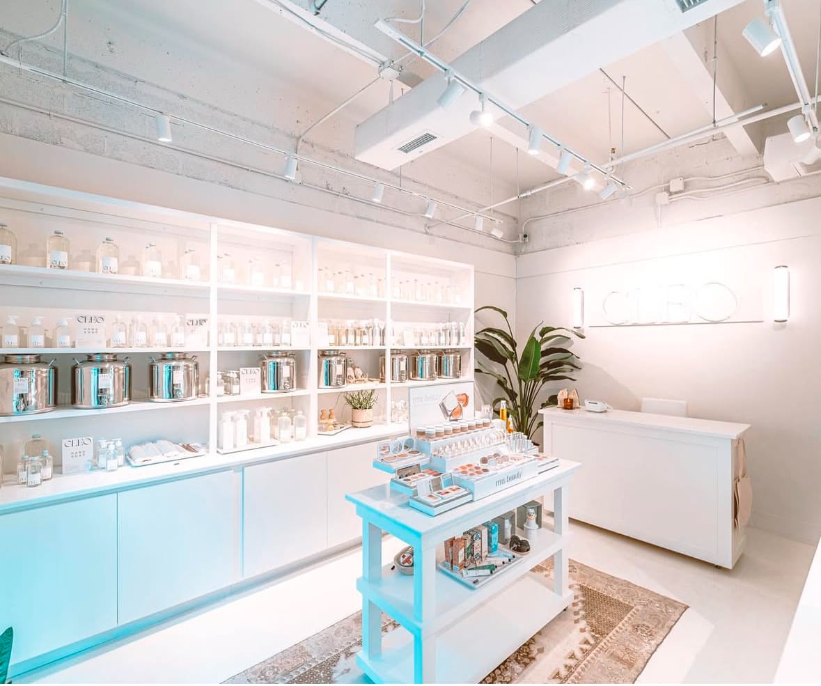 Bright, minimalist retail store interior with white shelves displaying various products. Plants add greenery, and a counter is positioned on the right. Ceiling has exposed ducts and lighting.