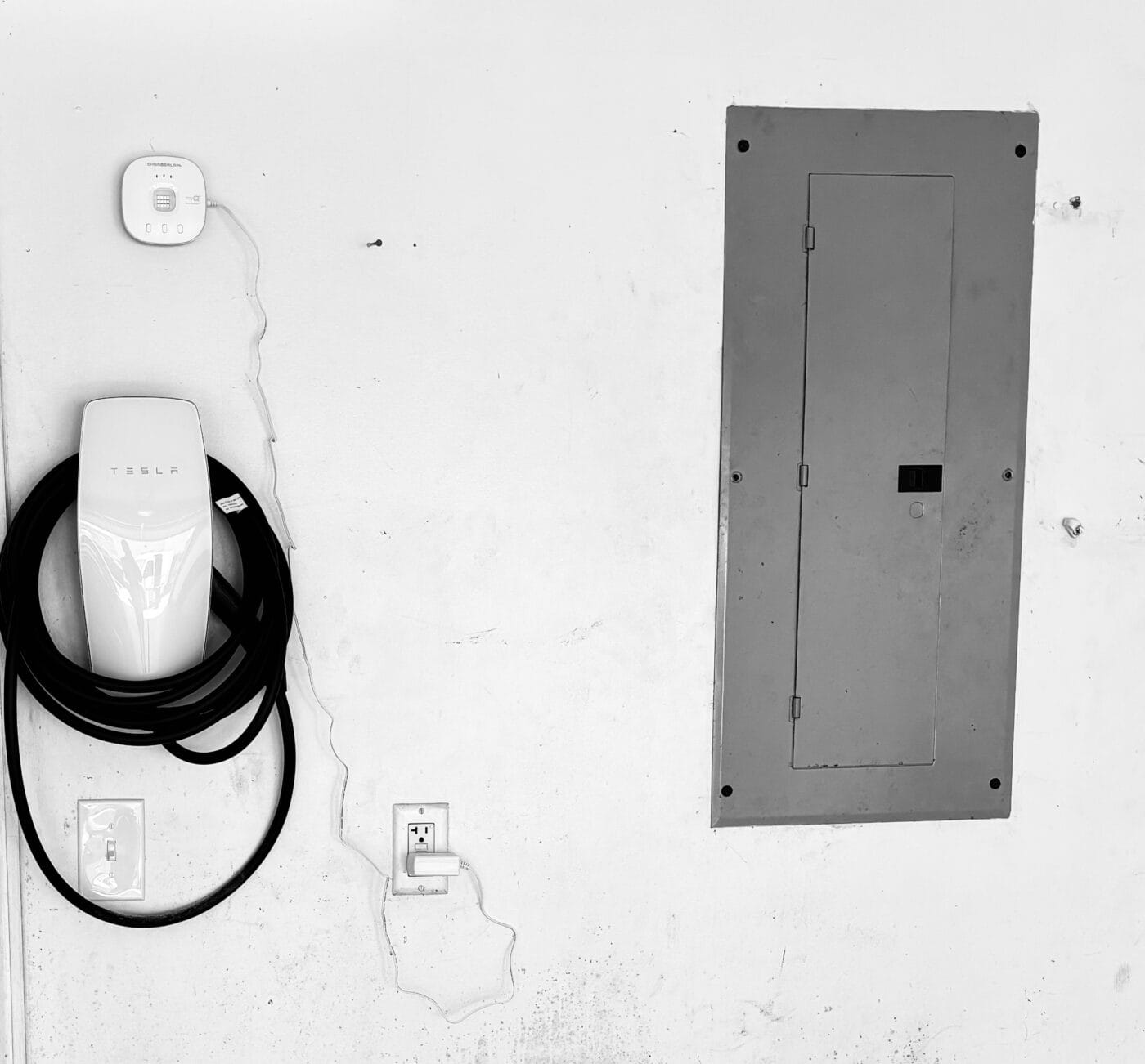 Wall-mounted Tesla charger with coiled cable next to an electrical panel and a thermostat on a white wall.