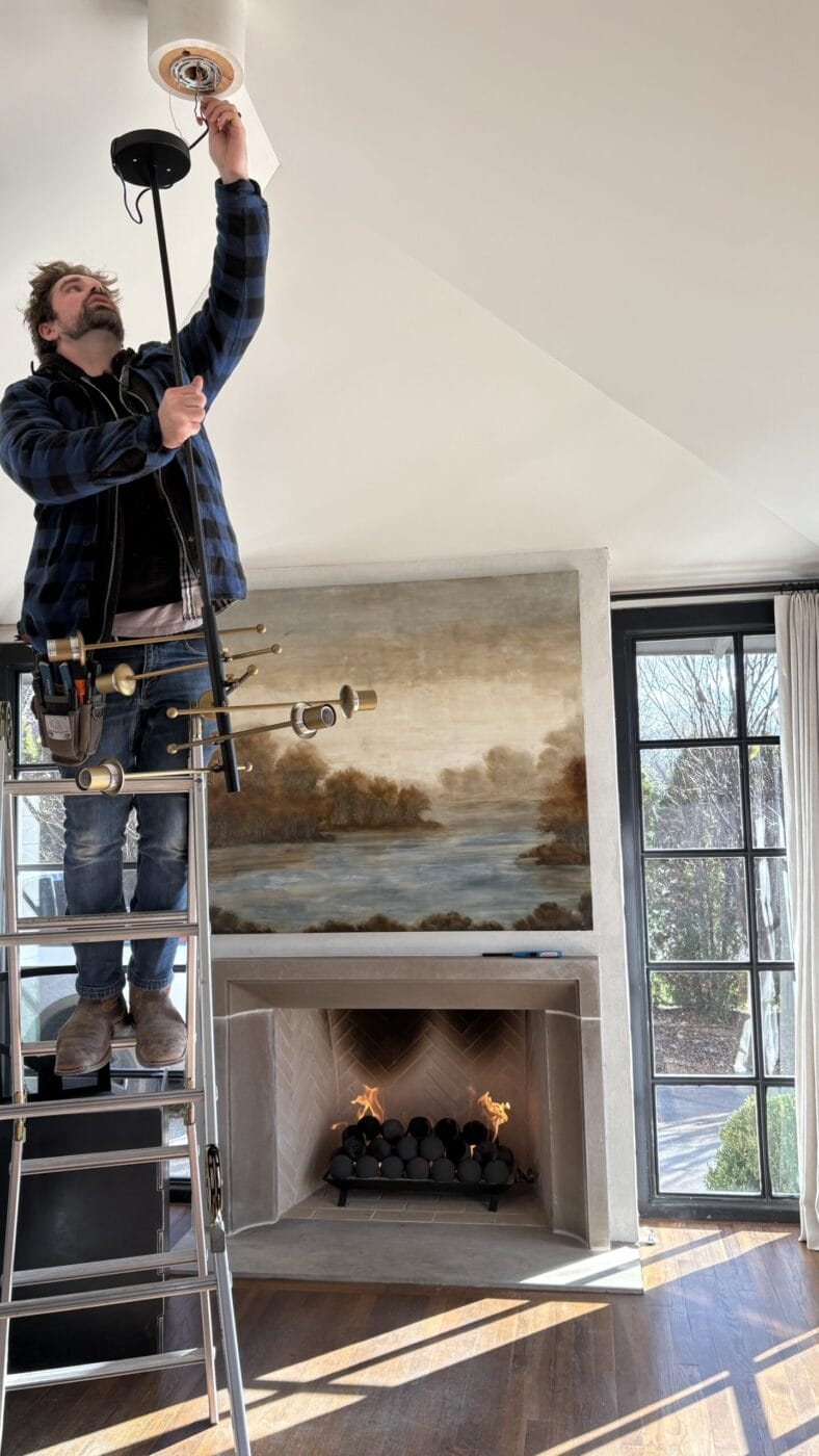 A person stands on a ladder installing a chandelier in a room with a fireplace, large painting, and floor-to-ceiling windows.