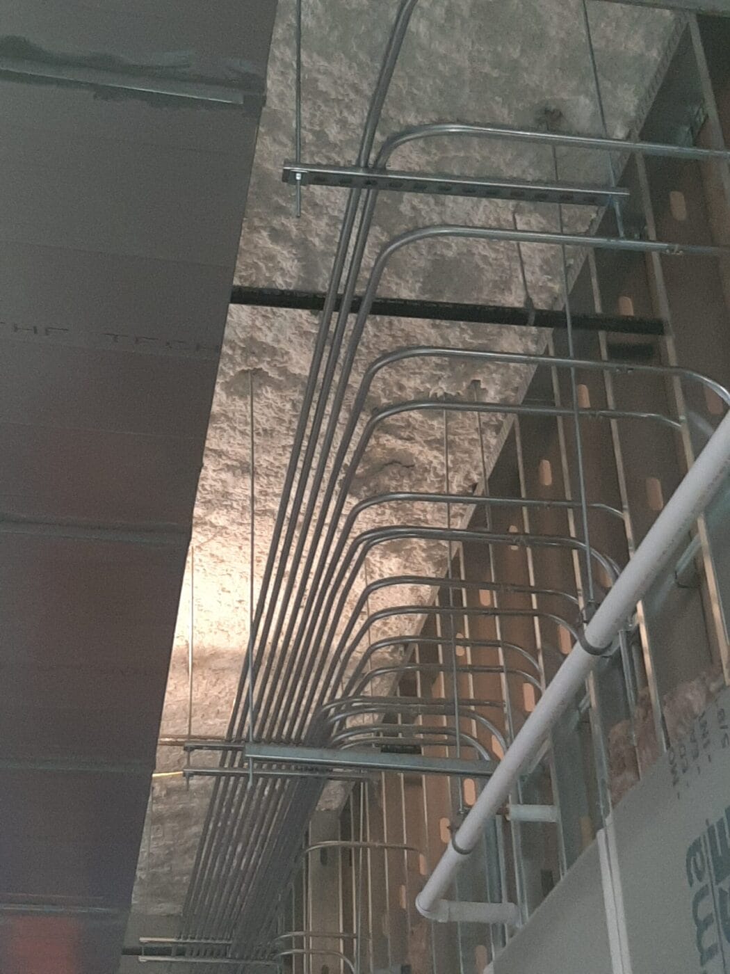 View of a ceiling with a series of curved metal pipes and tubing running parallel, with a textured concrete surface above.
